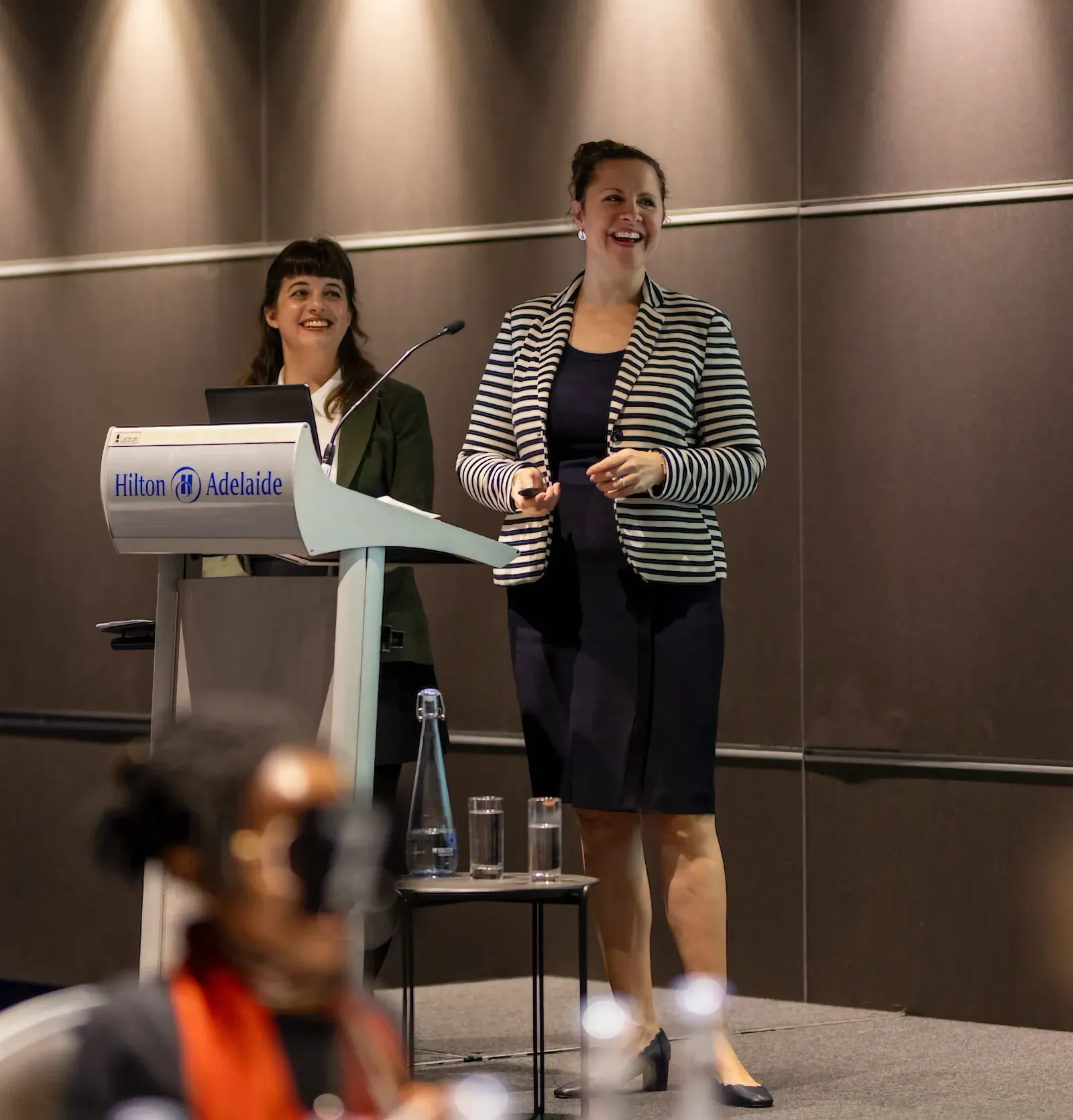 Two women presenting at a podium in a conference room, smiling as they address an audience.